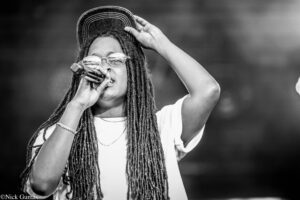 Black-and-white photo of a female singer with long dreadlocks, glasses, and a cap, singing into a handheld microphone on stage