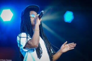 Singer performing on stage with a microphone, blue stage lights, and long dreadlocks; wearing a cap and round glasses.