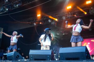 Three performers dance on a concert stage under orange spotlights, with large speakers and lighting rigs overhead.
