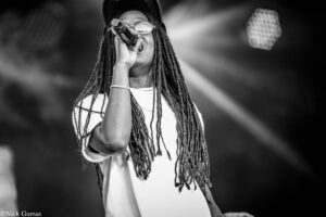 Black-and-white image of a singer with long dreadlocks, wearing a cap and glasses, singing into a microphone on stage.