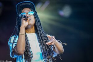 Performer singing into a microphone on stage, wearing a cap, round reflective sunglasses, and long dreadlocks.