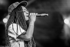 Black-and-white photo of a female singer with long dreadlocks, wearing a cap and glasses, singing into a microphone on stage.