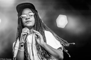 Black-and-white photo of a singer with long dreadlocks, wearing a cap and round glasses, holding a microphone on stage.