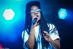 Close-up of a Black male singer with long dreadlocks, glasses, and a cap, singing into a microphone on a blue-lit stage.