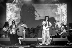 Live concert: central female singer with dreadlocks and glasses, mic in hand, flanked by guitarists on stage.