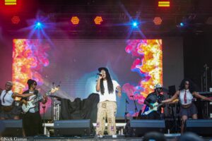 Lead performer center stage in a white shirt and patchwork pants, with band members around; fiery colorful LED backdrop and bright stage lights overhead.