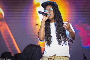 Female vocalist singing into a handheld microphone on stage, wearing a cap and white shirt with long dreadlocks.