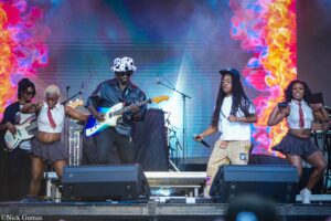 Group of musicians and dancers performing on a colorful stage with guitars and microphones, captured mid-performance at a concert.