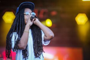 Person singing into a microphone on stage with long dreadlocks and round glasses, wearing a cap and white shirt.