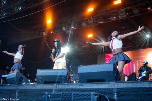 Stage performers in a lively concert: two dancers on either side with outstretched arms, rapper in cap center, bright orange stage lights in the background.
