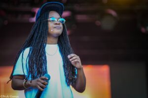 Female performer with long dreadlocks, round blue sunglasses, and a cap, holding a microphone on stage.