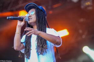 Female singer with dreadlocks wearing a blue cap and round sunglasses singing into a microphone on stage with warm lights in the background.
