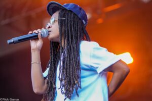Singer with long dreadlocks, blue cap and sunglasses singing into a handheld microphone on stage with warm orange lights behind.