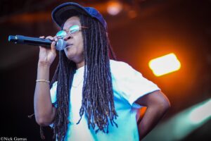 Female singer on stage, holding a microphone with long dreadlocks, wearing a blue cap and round sunglasses.