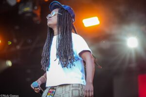 Musician with long dreadlocks wearing a blue cap and sunglasses, holding a microphone on stage with warm lights behind him/her.