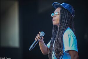 Person with long dreadlocks wearing a blue cap and reflective sunglasses sings into a handheld microphone on stage, wearing a light blue shirt.