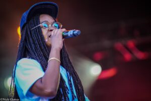 Female singer with dreadlocks, round glasses, and a blue cap singing into a microphone on stage.