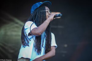 Singer with long dreadlocks and a blue cap, holding a microphone on stage against a dark background.