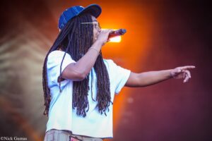 Female singer on stage, mic in hand, performing with long dreadlocks and a blue cap against warm orange backlighting.