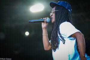 Female singer wearing a blue cap and glasses singing into a handheld microphone on stage.