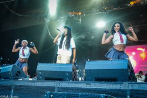 Three performers on a lit stage, center singer with mic in white shirt and blue cap, flanked by two dancers in skim outfits under bright stage lights.