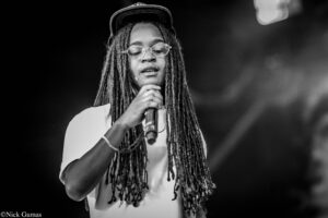 Singer performing on stage with long dreadlocks, round glasses, and a cap, holding a microphone close to face (black and white).