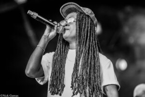 Female singer wearing a cap and round glasses, singing into a handheld microphone on stage (black-and-white).