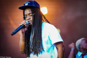 Singer with long dreadlocks, glasses, and a blue cap singing into a handheld microphone on stage.
