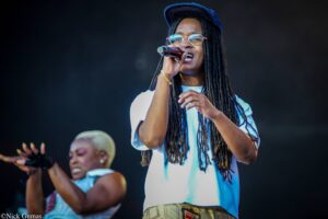 Female vocalist with dreadlocks and round glasses singing into a microphone on stage.