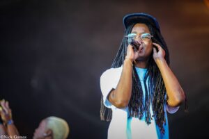 Female singer with glasses and long dreadlocks sings into a handheld microphone on stage, wearing a blue shirt and cap, bathed in stage light.