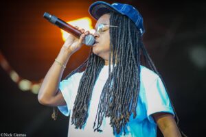 Performer singing into a handheld microphone on stage, wearing a blue cap and light blue shirt with long dreadlocks visible beyond the mic.