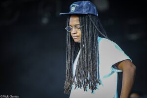 Portrait of a person with long dreadlocks, wearing a blue cap and round glasses, in a light blue shirt against a dark background.