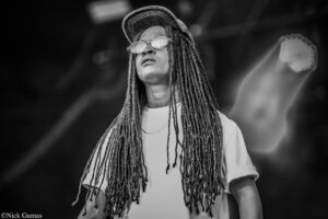 Black-and-white photo of a musician with long dreadlocks, a cap, and round sunglasses performing on stage, with a small star sticker on the cheek.