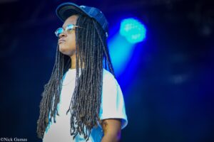 Person with long dreadlocks wearing a denim cap and round sunglasses looks upward, standing on a stage with blue lighting behind.