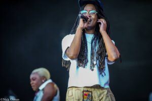 Female singer on stage singing into a microphone, wearing round glasses, a blue cap, and long dreadlocks.