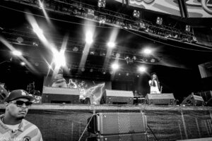 Black-and-white concert scene with performers on a lit stage, bright spotlights cutting through the air, and large speaker stacks in front of the stage.