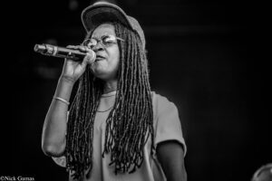 Black-and-white photo of a female singer with long dreadlocks, glasses, and a cap, singing into a handheld microphone on stage.