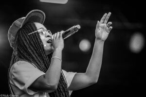 Black-and-white image of a woman singer with dreadlocks, wearing glasses and a cap, singing into a microphone with one hand raised.