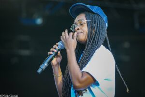 Female singer performing on stage, wearing a blue cap and glasses, holding a mic close to her mouth with long dreadlocks flowing behind her.