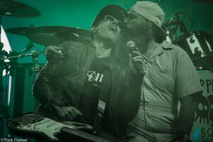Two musicians perform on stage: one sings into a microphone while playing a keyboard; the other leans in with a guitar and sunglasses under green stage lights.