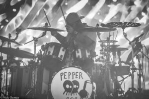 Black-and-white photo of a drummer wearing a hat behind a full drum kit on stage, with a bass drum displaying a Pepper logo and pepper graphics