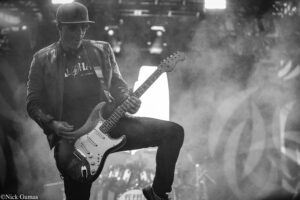 Guitarist wearing a leather jacket and sunglasses plays a white Stratocaster on a smoky stage.
