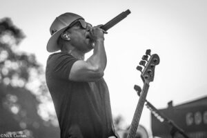 Male performer on stage singing into a handheld microphone, wearing a cap and sunglasses, with a guitar visible beside him.