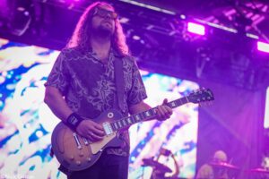 Guitarist with long hair and sunglasses playing a sunburst electric guitar on stage under purple lights.