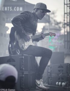 Guitarist wearing a cap and sunglasses, playing a white electric guitar on stage behind amplifiers.