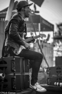 Guitarist in a leather jacket and cap plays an electric guitar on a stage, side profile with amps in the foreground.