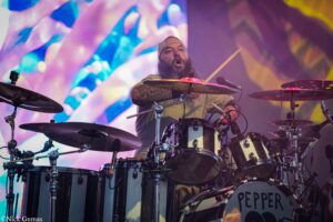 Drummer with a beard and backward cap playing a drum kit on a vibrant concert stage backdrop.