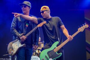 Two guitarists perform on a blue-lit stage—the left musician plays a white Strat-style guitar, the right musician plays a green bass. One points toward the audience as they perform.
