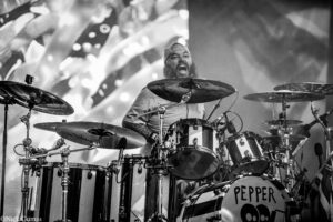 Bearded drummer in a cap, shouting with mouth open, surrounded by cymbals and a large drum kit on stage; bass drum reads 'PEPPER'.