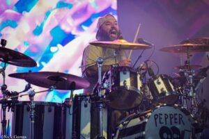 Drummer with long beard and backwards cap playing a drum kit on a brightly lit stage behind colorful lights.